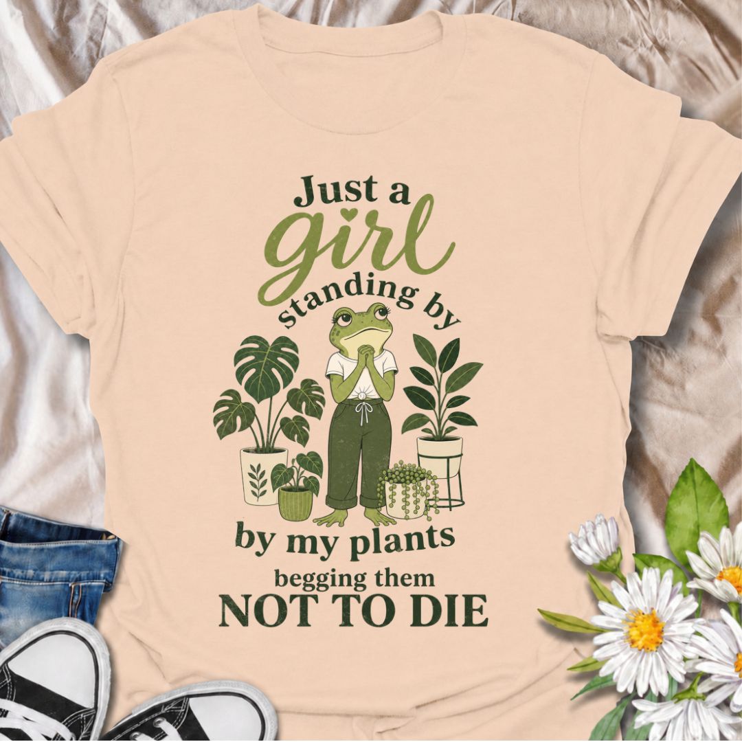 A funny plant T-shirt for anyone who spends far too much time encouraging their houseplants to stay alive. The phrase “Just a Girl Standing by My Plants Begging Them Not to Die” delivers relatable, self-deprecating humor for plant parents and indoor gardeners. Featuring a frog character surrounded by potted plants, the design feels playful, niche, and easy to wear. It makes a great gift for houseplant lovers, plant collectors, and anyone who has ever stared anxiously at a struggling leaf. Perfect for nurser