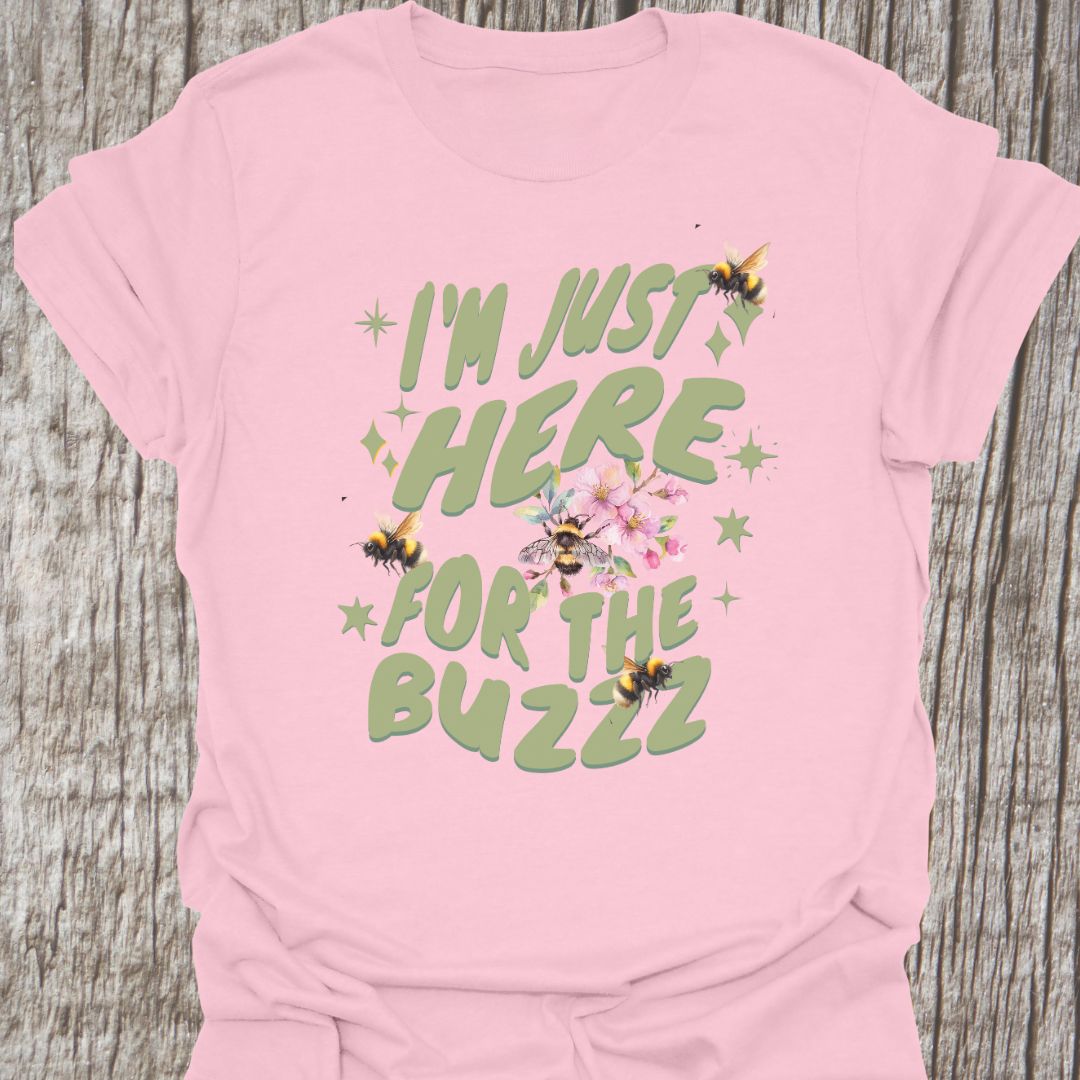 I am Just Here For The Buzz light pink T-shirt, featuring a green text design adorned with bees and flowers for decoration