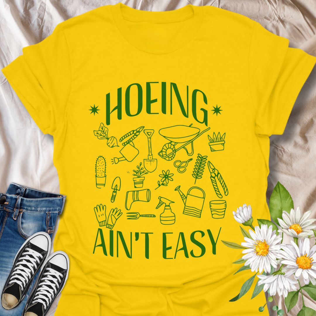 Funny gardening t-shirt with cute garden tools and “Hoeing Ain’t Easy” slogan. Perfect gift for plant lovers, gardeners, or anyone with a green thumb and a sense of humor. Daisy