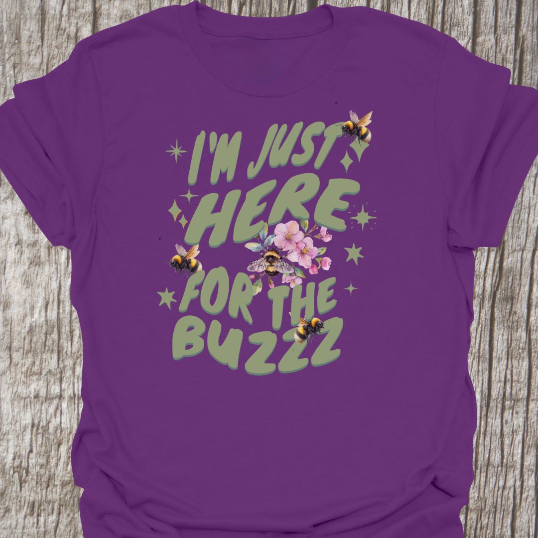 I am Just Here For The Buzz purple T-shirt, featuring a green text design adorned with bees and flowers for decoration