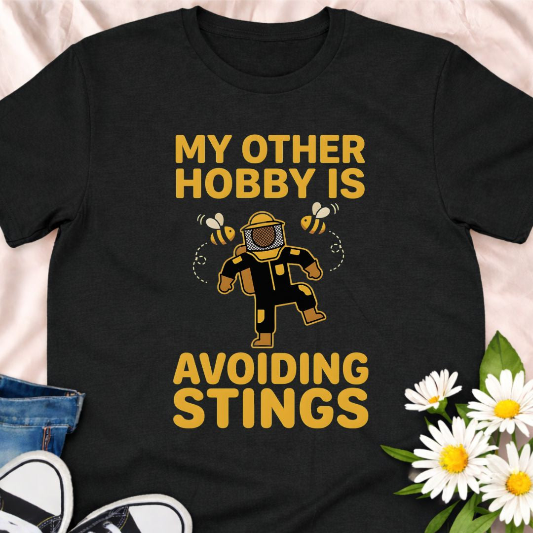 My other hobby is avoiding stings. A funny beekeeper shirt featuring a playful bee graphic, perfect for beekeepers, honey lovers, and anyone obsessed with bees. This soft unisex tee makes a great gift for apiarists, gardeners, and pollinator supporters.