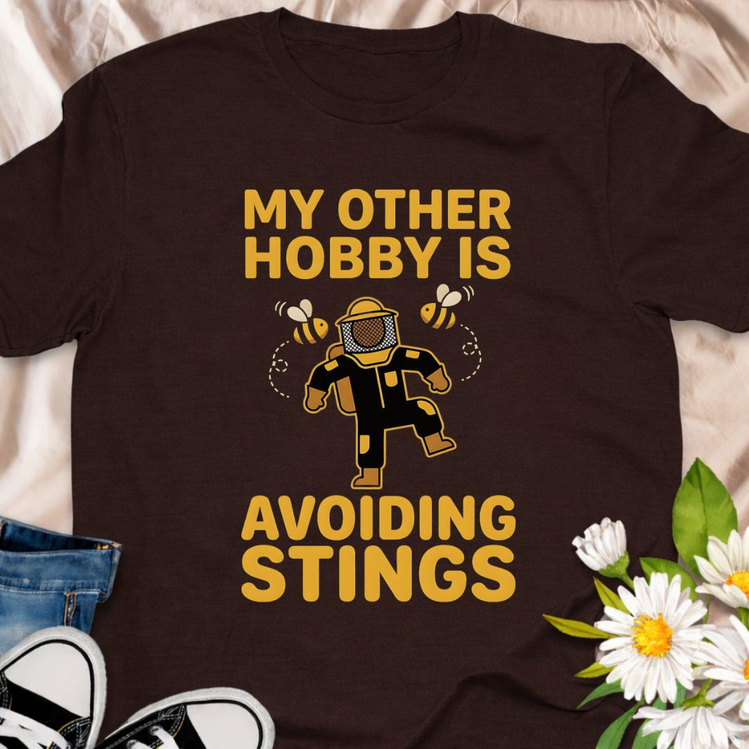 My other hobby is avoiding stings. A funny beekeeper shirt featuring a playful bee graphic, perfect for beekeepers, honey lovers, and anyone obsessed with bees. This soft unisex tee makes a great gift for apiarists, gardeners, and pollinator supporters.