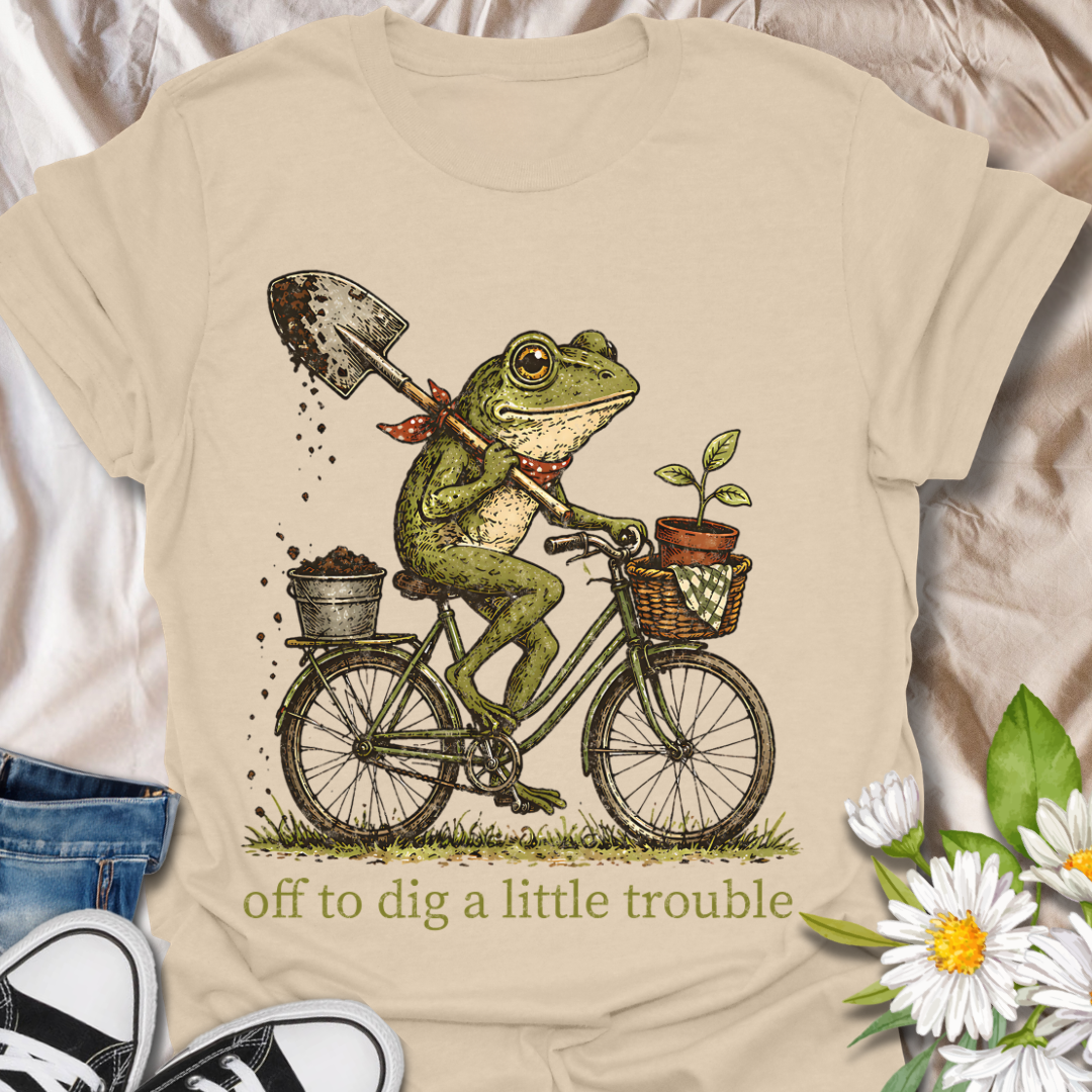 Funny gardening t-shirt featuring a frog riding a bike with a shovel and plant pot, paired with the phrase “Off to Dig a Little Trouble.” Perfect for gardeners, plant lovers, frog enthusiasts, and anyone who enjoys playful garden humor.