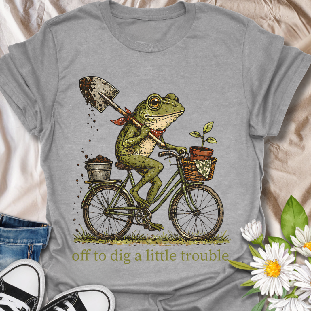 Funny gardening t-shirt featuring a frog riding a bike with a shovel and plant pot, paired with the phrase “Off to Dig a Little Trouble.” Perfect for gardeners, plant lovers, frog enthusiasts, and anyone who enjoys playful garden humor.