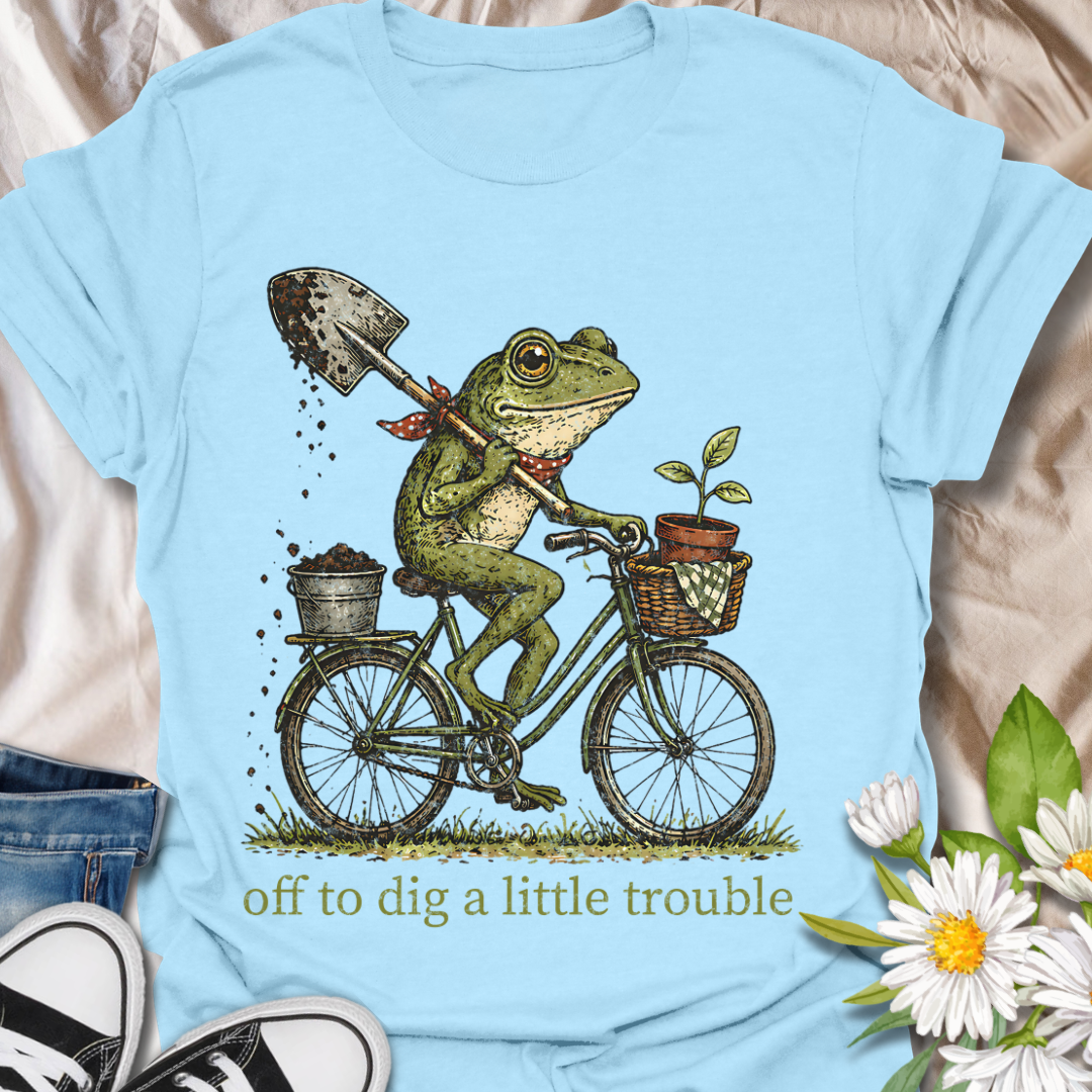 Funny gardening t-shirt featuring a frog riding a bike with a shovel and plant pot, paired with the phrase “Off to Dig a Little Trouble.” Perfect for gardeners, plant lovers, frog enthusiasts, and anyone who enjoys playful garden humor.