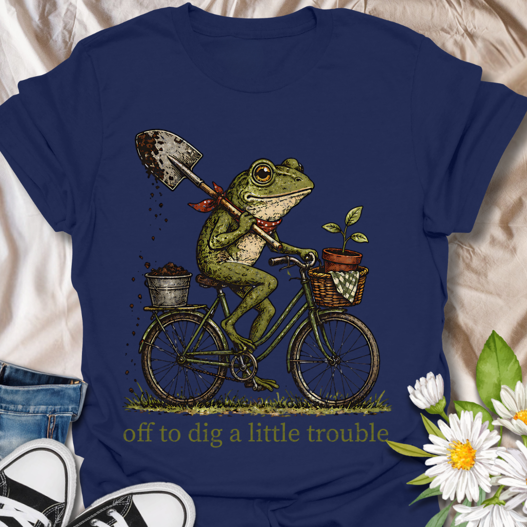 Funny gardening t-shirt featuring a frog riding a bike with a shovel and plant pot, paired with the phrase “Off to Dig a Little Trouble.” Perfect for gardeners, plant lovers, frog enthusiasts, and anyone who enjoys playful garden humor.