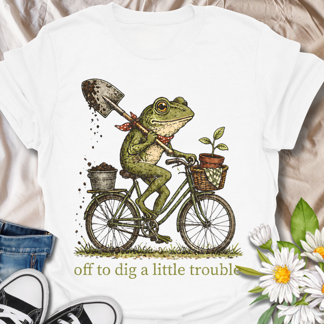 Funny gardening t-shirt featuring a frog riding a bike with a shovel and plant pot, paired with the phrase “Off to Dig a Little Trouble.” Perfect for gardeners, plant lovers, frog enthusiasts, and anyone who enjoys playful garden humor.