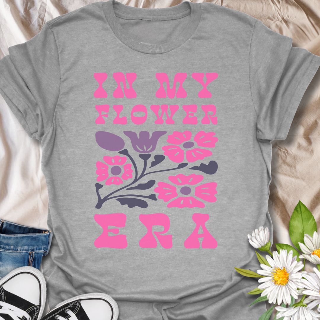 This In My Flower Era t-shirt is made for flower lovers who are fully embracing their blooming, botanical, garden-girl phase. Featuring bold retro pink lettering with a simple floral graphic in soft purple and navy tones, this design has a playful vintage-inspired look that feels cute, bright, and easy to wear. Perfect for gardeners, flower fans, plant lovers, cottagecore style lovers, botanical art fans, and anyone who is proudly in their flower era.