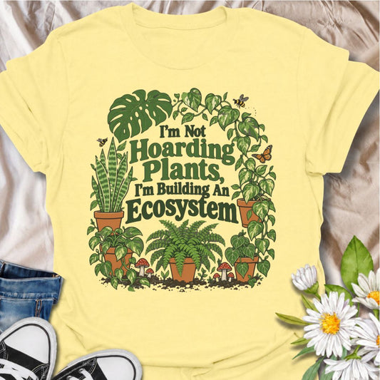 A funny plant T-shirt for anyone whose houseplant collection has grown well beyond “just a few plants.” The phrase “I’m Not Hoarding Plants, I’m Building an Ecosystem” turns plant collecting into a perfectly reasonable lifestyle choice with playful, relatable humor. Surrounded by monstera, pothos, snake plants, ferns, mushrooms, and pollinators, the design feels lush without losing readability. It makes a great gift for plant parents, indoor gardeners, houseplant collectors, and anyone proudly creating thei