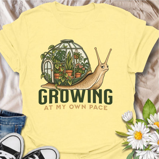A whimsical plant T-shirt for anyone who believes growth does not need to be rushed. Featuring a laid-back snail carrying a tiny greenhouse filled with lush houseplants, this design pairs the phrase “Growing at My Own Pace” with a calm, slightly quirky botanical illustration. It is perfect for plant lovers, gardeners, slow-living fans, and anyone who enjoys a gentle reminder to take life one step at a time. The detailed artwork gives it a distinctive storybook feel while still staying clearly rooted in the 