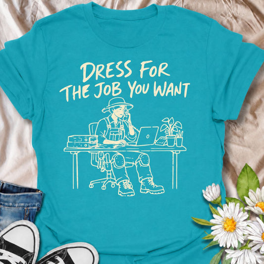 A funny gardening T-shirt for anyone who would rather be dressed for the garden than stuck behind a desk. The phrase “Dress for the Job You Want” pairs with a gardener in full work gear sitting in an office setting for a dry, absurd bit of garden humor. Featuring overalls, knee pads, work boots, tools, a laptop, and desk accessories, the design is perfect for gardeners who know exactly where they would rather be. It makes a great gift for professional gardeners, backyard growers, plant lovers, and anyone dr