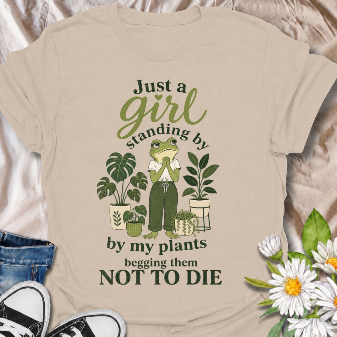 A funny plant T-shirt for anyone who spends far too much time encouraging their houseplants to stay alive. The phrase “Just a Girl Standing by My Plants Begging Them Not to Die” delivers relatable, self-deprecating humor for plant parents and indoor gardeners. Featuring a frog character surrounded by potted plants, the design feels playful, niche, and easy to wear. It makes a great gift for houseplant lovers, plant collectors, and anyone who has ever stared anxiously at a struggling leaf. Perfect for nurser