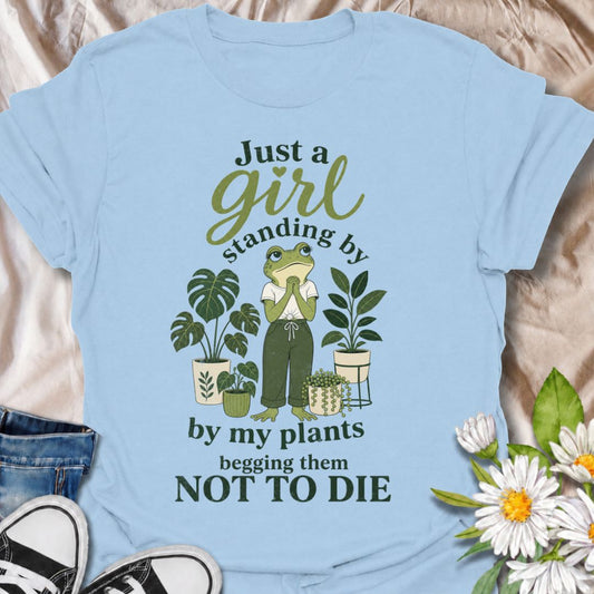 A funny plant T-shirt for anyone who spends far too much time encouraging their houseplants to stay alive. The phrase “Just a Girl Standing by My Plants Begging Them Not to Die” delivers relatable, self-deprecating humor for plant parents and indoor gardeners. Featuring a frog character surrounded by potted plants, the design feels playful, niche, and easy to wear. It makes a great gift for houseplant lovers, plant collectors, and anyone who has ever stared anxiously at a struggling leaf. Perfect for nurser