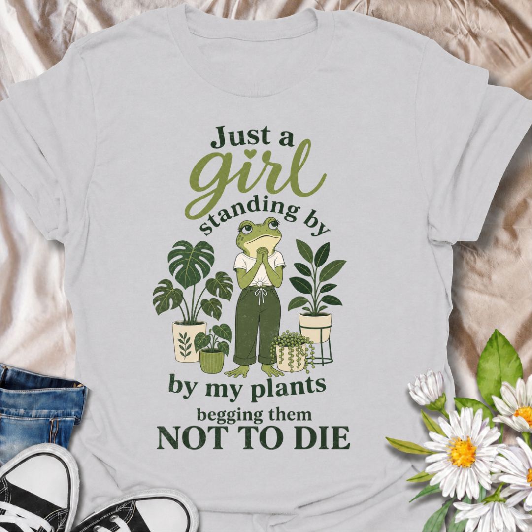 A funny plant T-shirt for anyone who spends far too much time encouraging their houseplants to stay alive. The phrase “Just a Girl Standing by My Plants Begging Them Not to Die” delivers relatable, self-deprecating humor for plant parents and indoor gardeners. Featuring a frog character surrounded by potted plants, the design feels playful, niche, and easy to wear. It makes a great gift for houseplant lovers, plant collectors, and anyone who has ever stared anxiously at a struggling leaf. Perfect for nurser