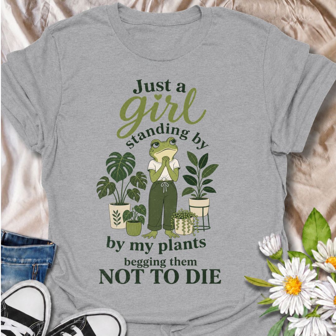 A funny plant T-shirt for anyone who spends far too much time encouraging their houseplants to stay alive. The phrase “Just a Girl Standing by My Plants Begging Them Not to Die” delivers relatable, self-deprecating humor for plant parents and indoor gardeners. Featuring a frog character surrounded by potted plants, the design feels playful, niche, and easy to wear. It makes a great gift for houseplant lovers, plant collectors, and anyone who has ever stared anxiously at a struggling leaf. Perfect for nurser