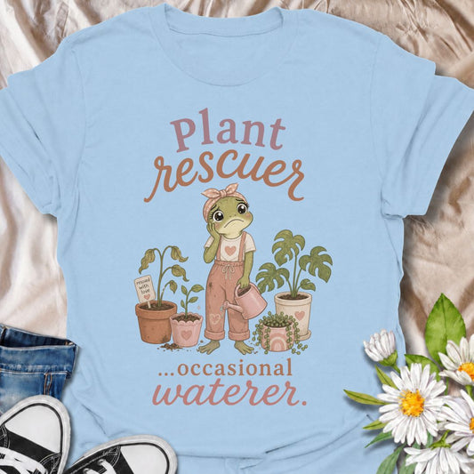 A funny plant T-shirt for anyone who cannot resist trying to save a struggling houseplant. The phrase “Plant Rescuer… Occasional Waterer” pairs with a sweet frog character, wilted plants, watering can, and potted greenery for playful, self-aware plant-parent humor. It is perfect for houseplant lovers, indoor gardeners, and anyone who brings home sad plants with every intention of nursing them back to health. The softer pastel artwork gives the design a warm, feminine feel while keeping the joke clear and re