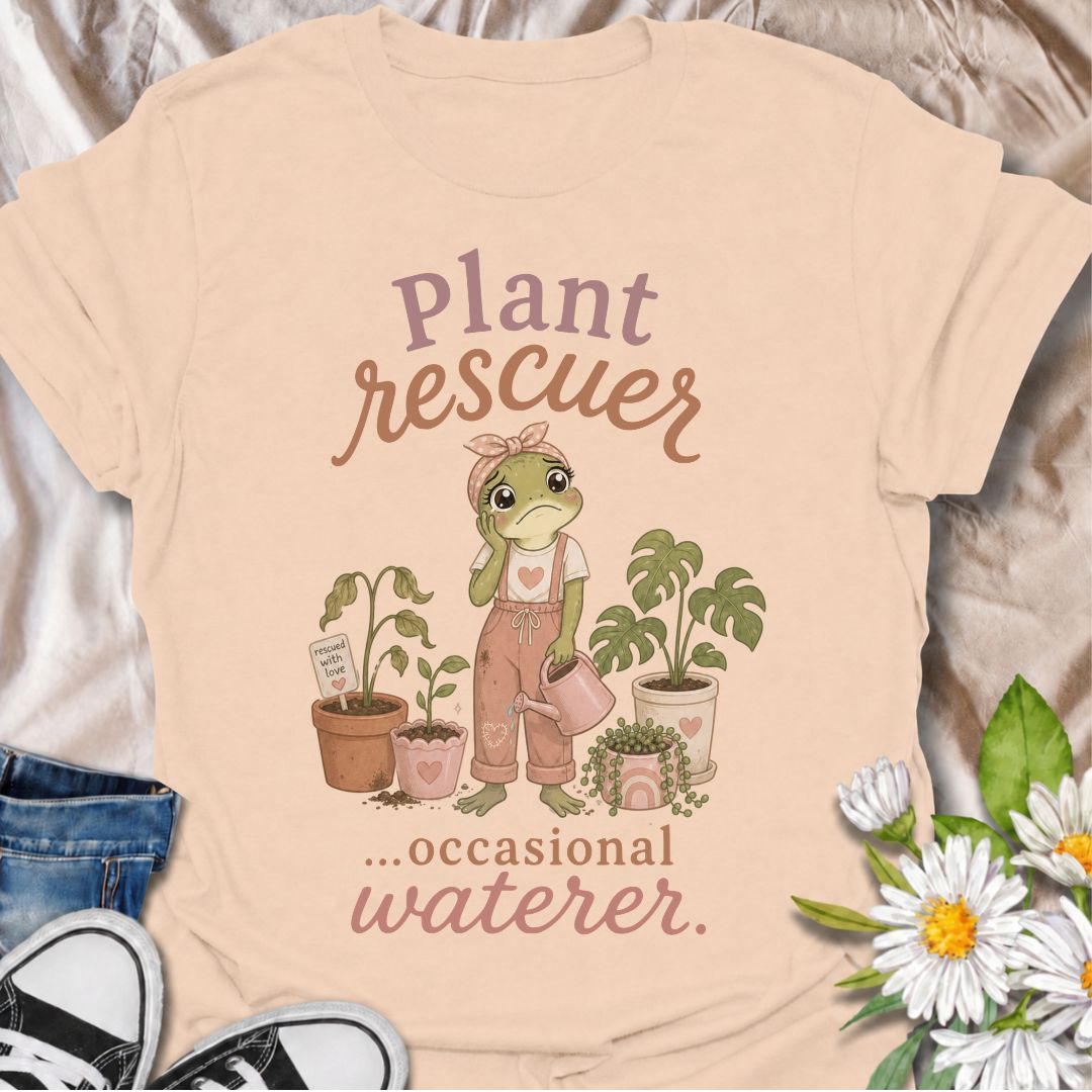 A funny plant T-shirt for anyone who cannot resist trying to save a struggling houseplant. The phrase “Plant Rescuer… Occasional Waterer” pairs with a sweet frog character, wilted plants, watering can, and potted greenery for playful, self-aware plant-parent humor. It is perfect for houseplant lovers, indoor gardeners, and anyone who brings home sad plants with every intention of nursing them back to health. The softer pastel artwork gives the design a warm, feminine feel while keeping the joke clear and re