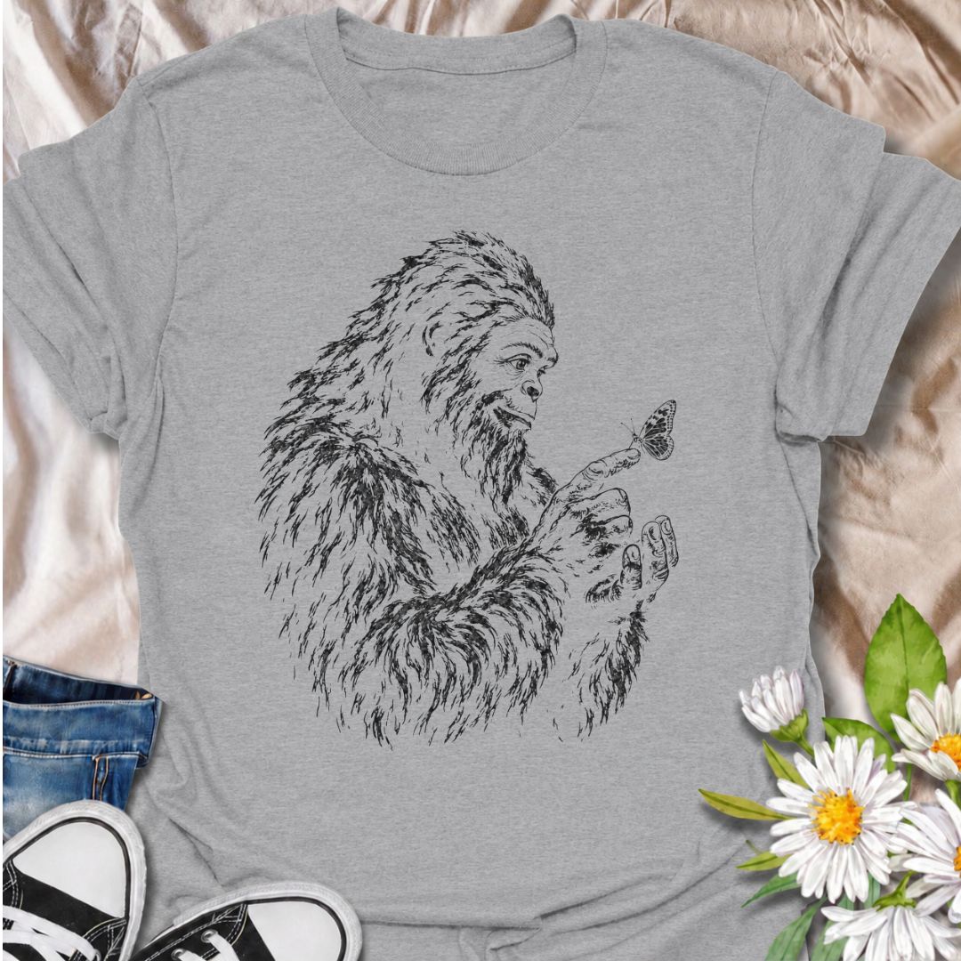 A gentle Bigfoot T-shirt for nature lovers who appreciate the quieter side of the wilderness. Featuring a hand-drawn Bigfoot carefully watching a butterfly resting on his fingertip, this design combines woodland folklore with a soft, unexpected moment of curiosity. It is perfect for Bigfoot fans, gardeners, pollinator lovers, hikers, and anyone drawn to whimsical nature-inspired artwork. The monochrome sketch style gives the design a vintage field-guide feel without becoming overly busy. A distinctive gift 
