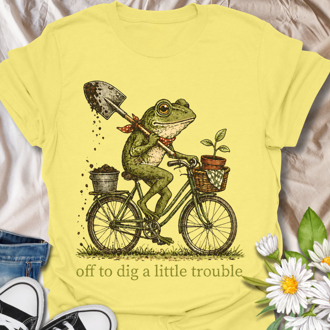 Funny gardening t-shirt featuring a frog riding a bike with a shovel and plant pot, paired with the phrase “Off to Dig a Little Trouble.” Perfect for gardeners, plant lovers, frog enthusiasts, and anyone who enjoys playful garden humor.