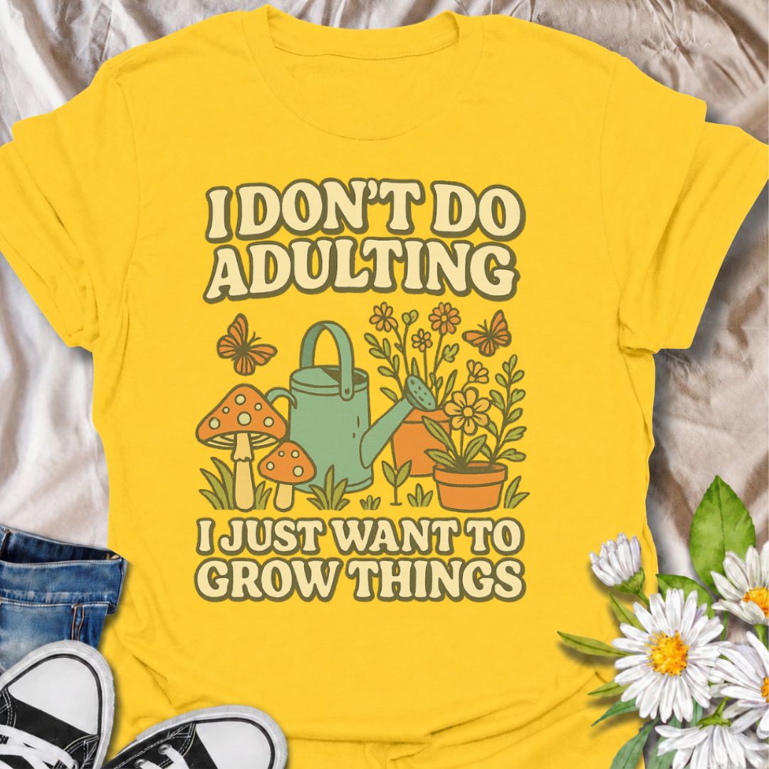 A bright yellow graphic t-shirt laid flat on a light beige fabric background, styled with blue jeans, black sneakers, and white daisies. The t-shirt features a retro-style graphic with the text 'I DON'T DO ADULTING I JUST WANT TO GROW THINGS' and an illustration of a green watering can, mushrooms, flowers, and butterflies.