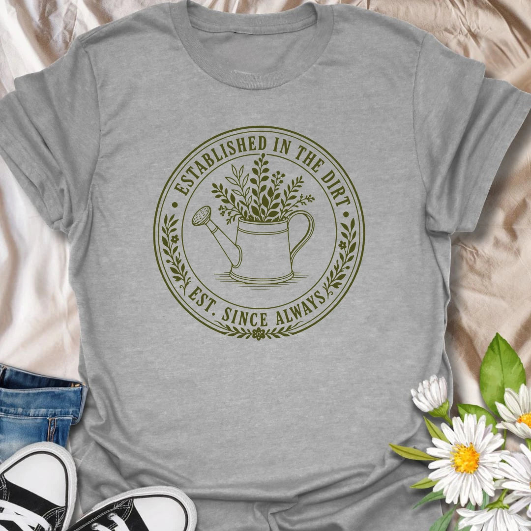 This Established In The Dirt t-shirt is made for gardeners who have always felt happiest with soil on their hands and something green to grow. Featuring a vintage-style circular badge with a watering can, leafy plant details, and the phrase Est. Since Always, this design has a simple botanical look that feels classic, earthy, and easy to wear. Perfect for gardeners, plant lovers, backyard growers, flower fans, vegetable gardeners, and anyone whose happy place is firmly rooted in the dirt.