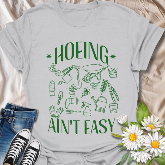 Funny gardening t-shirt with cute garden tools and “Hoeing Ain’t Easy” slogan. Perfect gift for plant lovers, gardeners, or anyone with a green thumb and a sense of humor