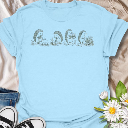 This Gardening Hedgehogs t-shirt is made for gardeners, nature lovers, and woodland animal fans who enjoy sweet vintage-style artwork. Featuring four charming hedgehogs watering, digging, carrying plants, and pushing a flower-filled wheelbarrow, this design has a gentle cottagecore garden feel. Perfect for plant lovers, gardeners, hedgehog fans, wildlife lovers, botanical art fans, and anyone who loves soft nature-inspired designs.