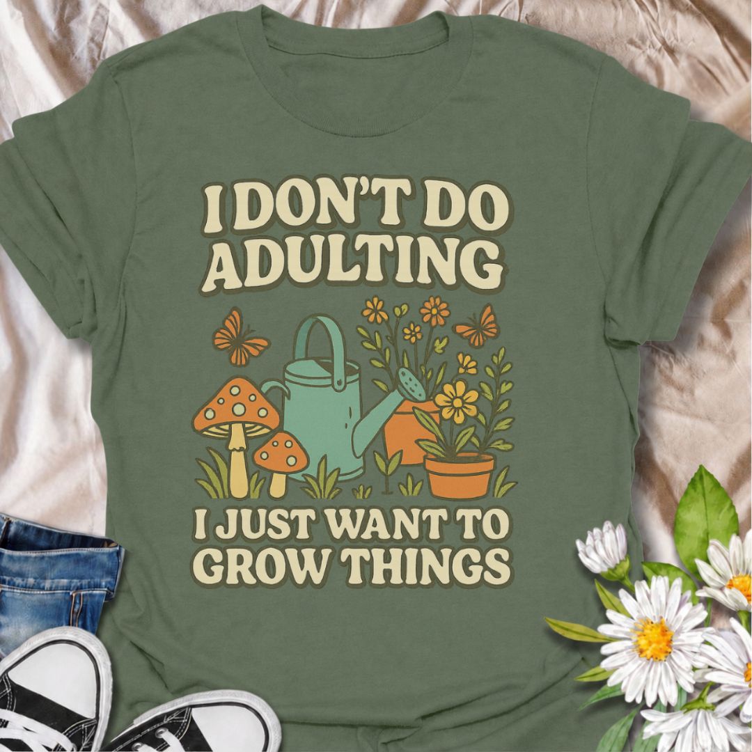A military green graphic t-shirt laid flat on a pink satin fabric background, styled with blue jeans, black sneakers, and white daisies. The t-shirt features a retro-style graphic with the text 'I DON'T DO ADULTING I JUST WANT TO GROW THINGS' in beige, accompanied by illustrations of a green watering can, orange mushrooms, yellow flowers, and butterflies.