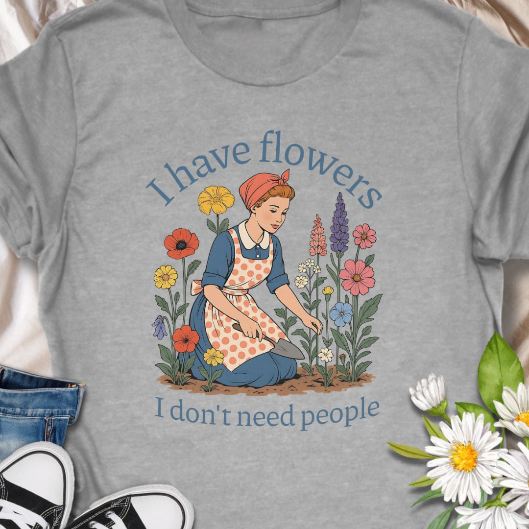 Express your introverted, plant-loving side with this charming floral t-shirt. Perfect for gardeners, plant moms, or anyone who finds peace in flowers over people — it's a blooming statement of self-care.  This tee pairs vintage garden charm with a wry sense of independence. A retro-style gardener tends bright, cottage blooms while the caption reads “I have flowers / I don’t need people.” 